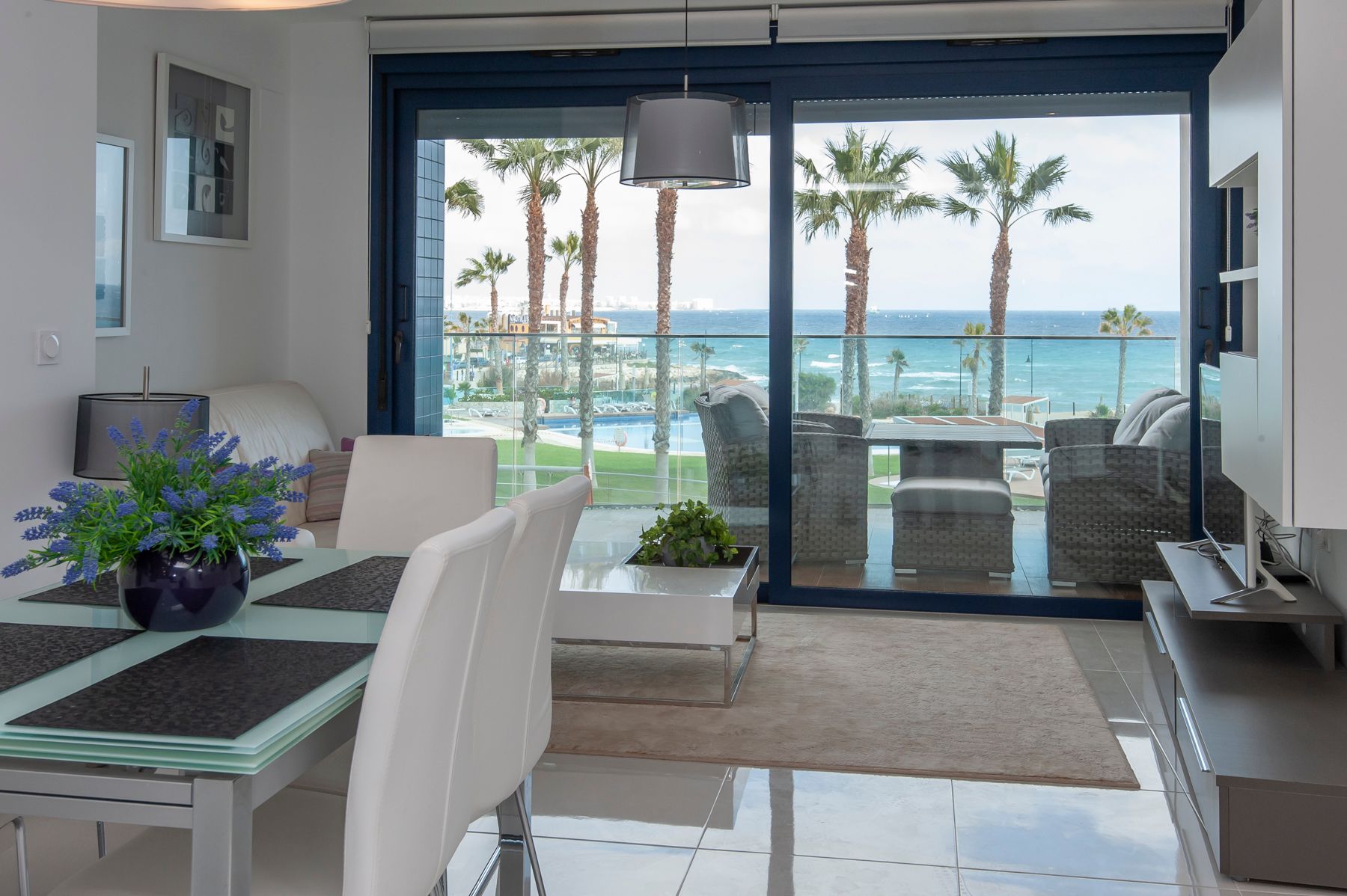 En stue med spisestuebord og utsikt til balkong og strand med palmer