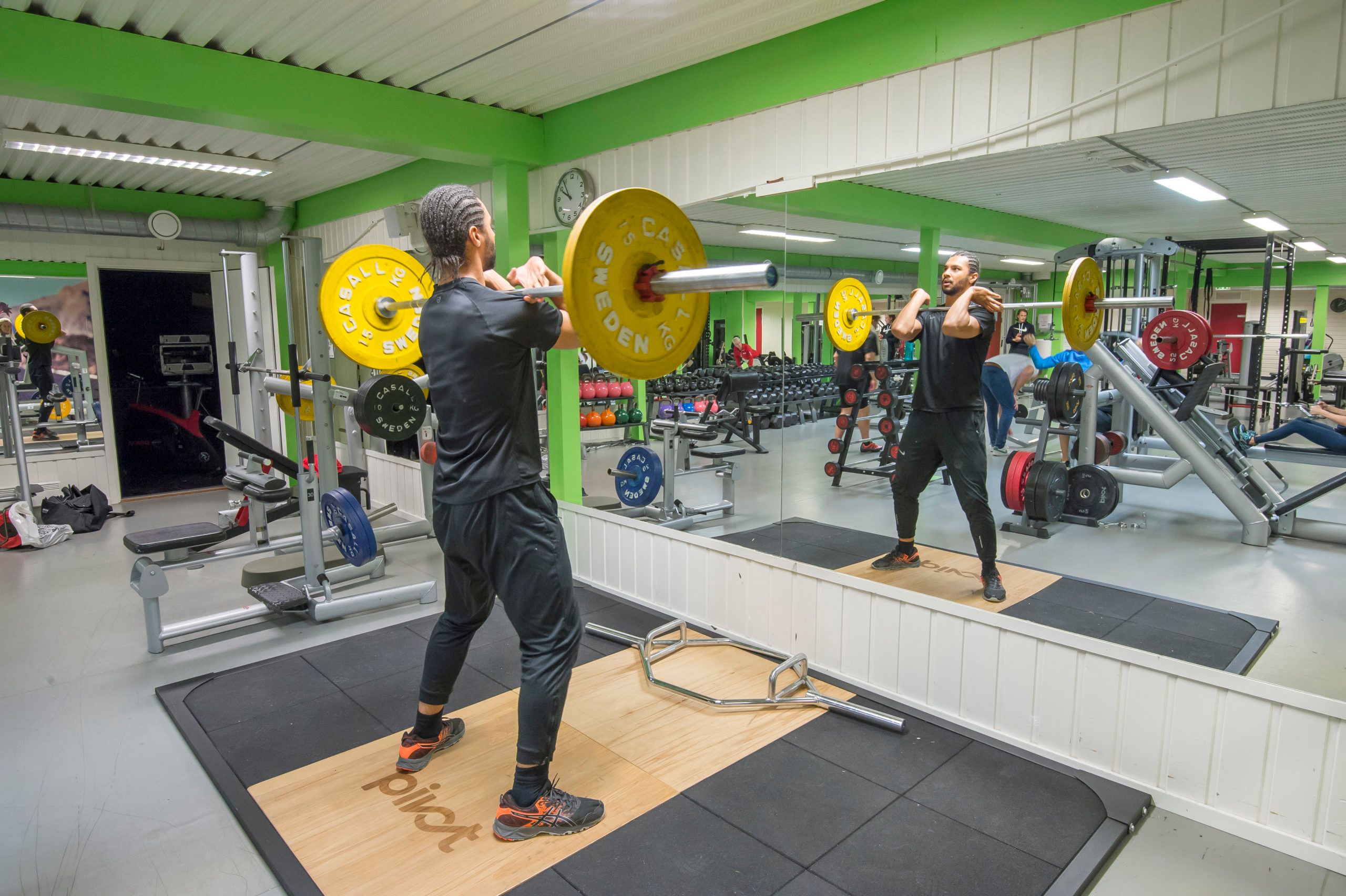 En mann som holder en vektstang på brystet foran et speil i et treningsstudio. Vektstand med en gul vekt på hver side