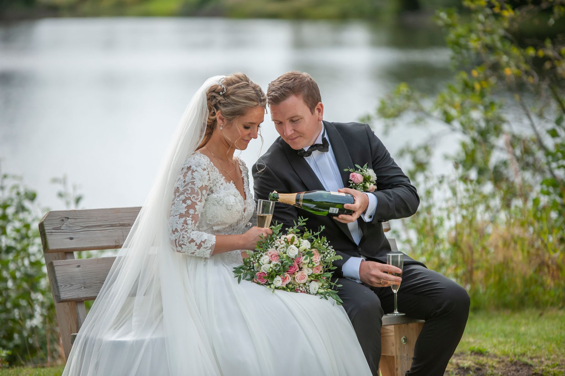 Brudepar som sitter på en benk, brudgommen skjenker et glass med sprudlende til bruden, med vann i bakgrunnen