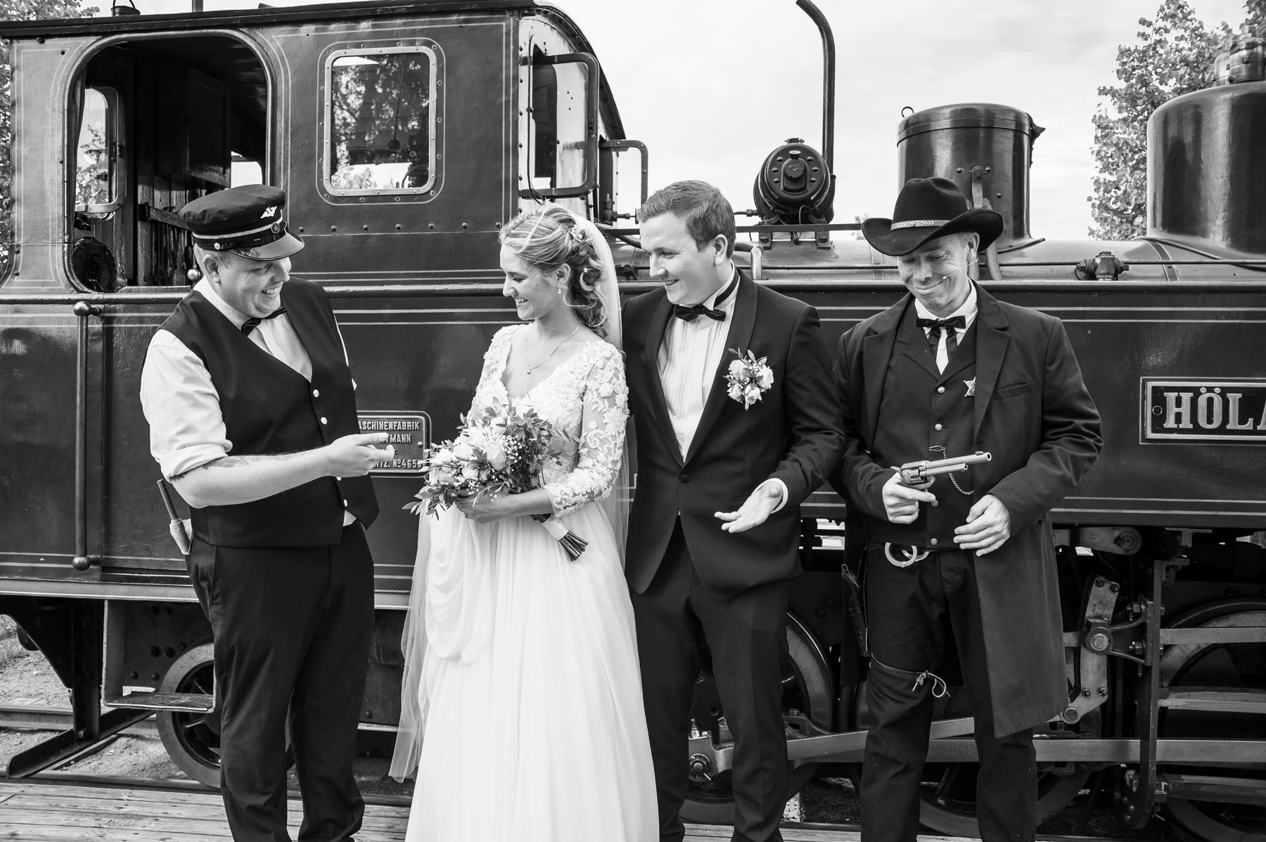 Et brudepar foran et damplokomotiv, flankert av lokfører og togrøver
